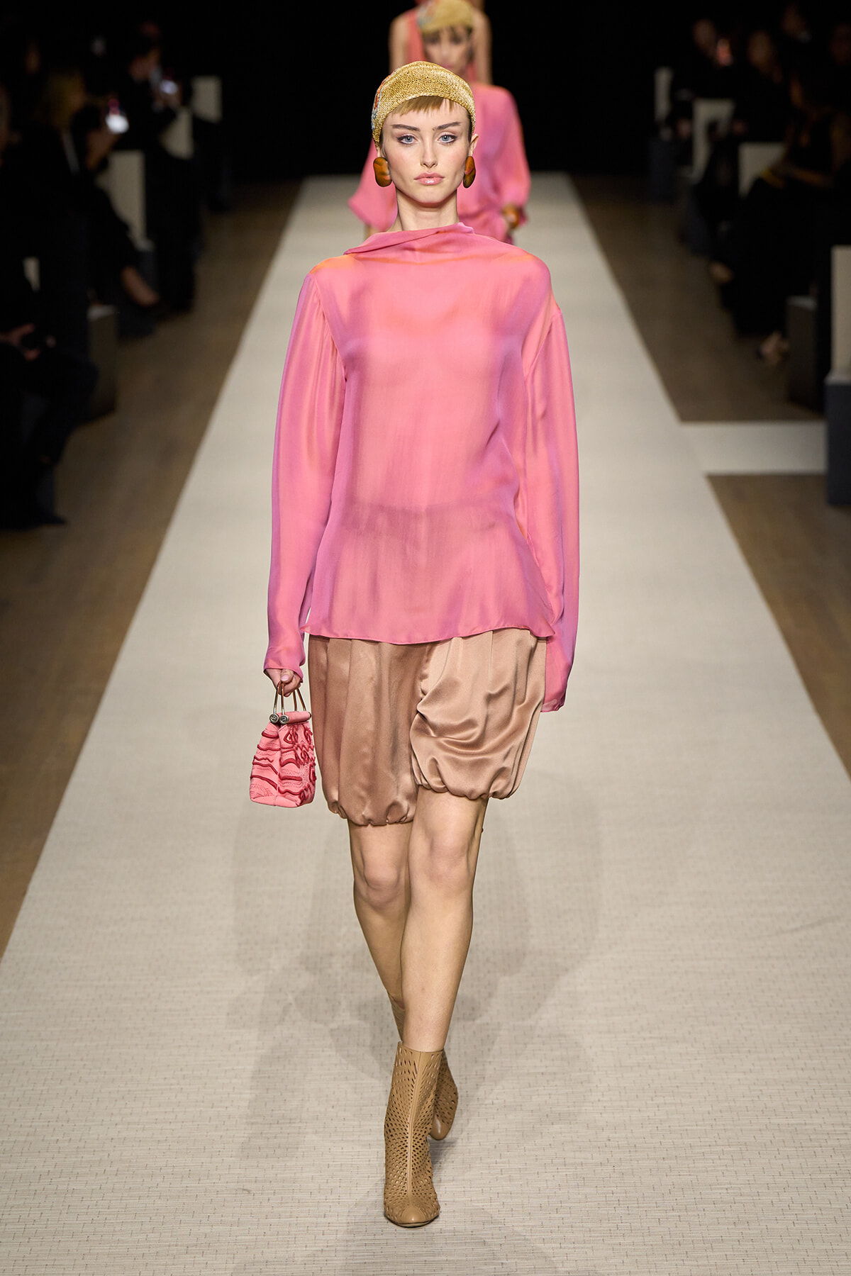Runway model in a pink sheer blouse with a high neck, paired with bronze satin bubble shorts, tan perforated ankle boots, and a pink mini handbag, wearing a gold headwrap.