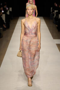 Model walks the runway in a pink marbled spaghetti-strap dress with a V-neck and a wide waist sash, carrying a small beige clutch and wearing a gold headwrap and earrings