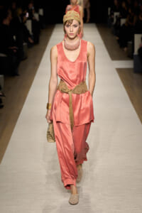 Model on a runway wearing coral satin sleeveless top and wide-leg pants with a knotted beige belt, head wrap, and statement necklace.