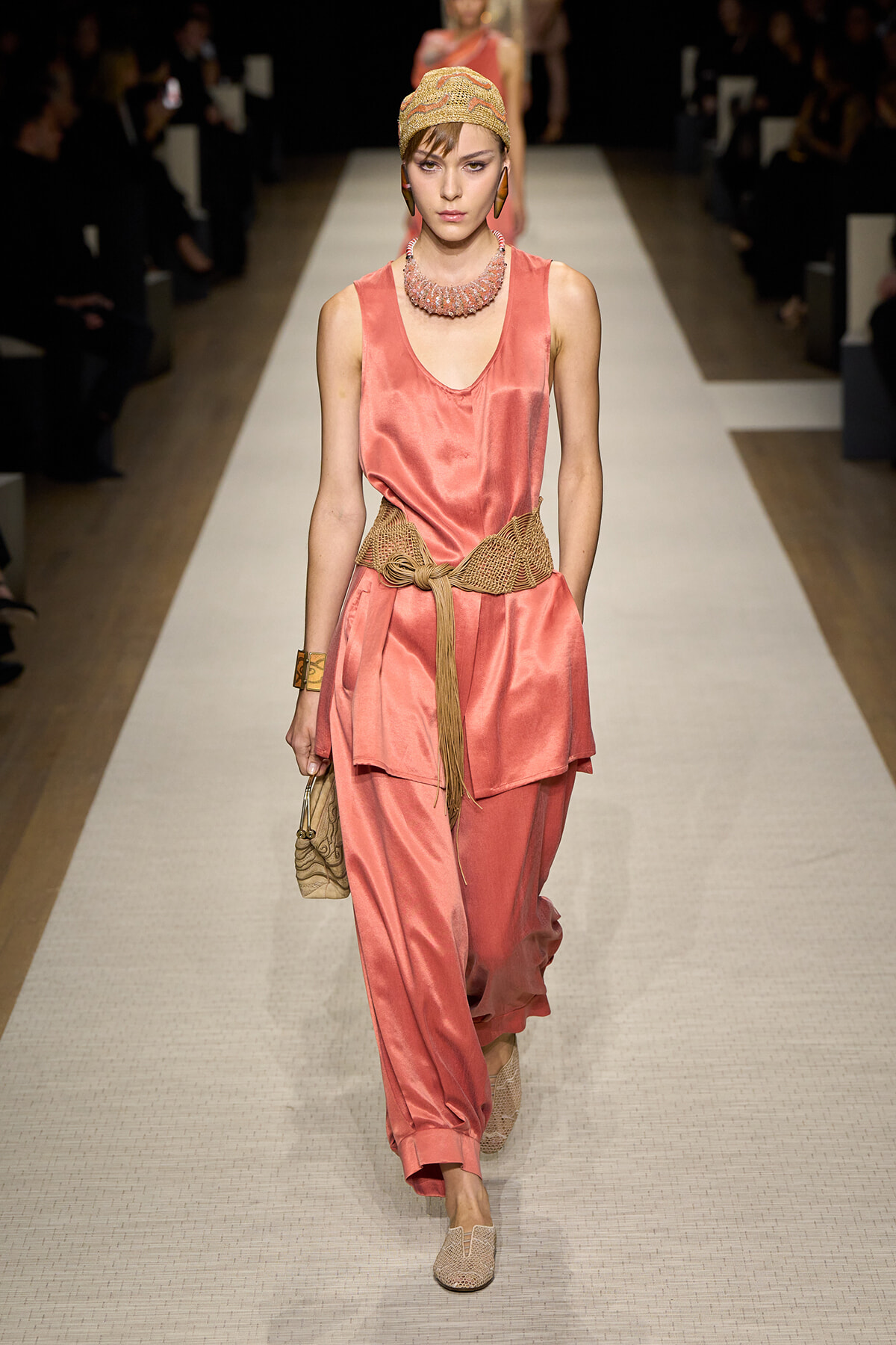 Model on a runway wearing coral satin sleeveless top and wide-leg pants with a knotted beige belt, head wrap, and statement necklace.
