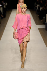 Model walking a runway in a pink draped dress with a gathered waist, beige shorts, tan perforated ankle boots, a gold headwrap, and a pink clutch.