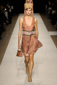Runway model walking the catwalk in a pink-brown patterned sleeveless dress with a multicolor waist stripe, headscarf, beige perforated boots, and a small brown handbag.