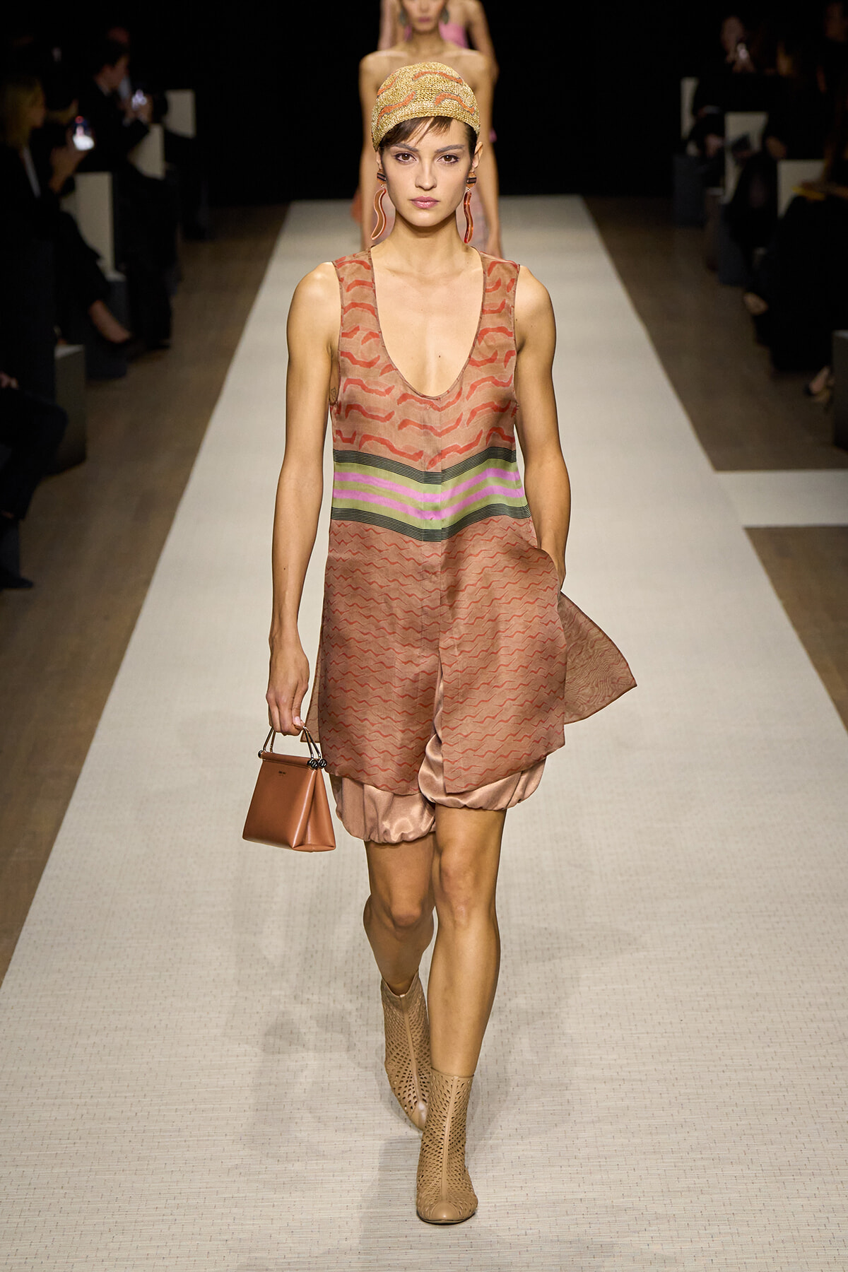 Runway model walking the catwalk in a pink-brown patterned sleeveless dress with a multicolor waist stripe, headscarf, beige perforated boots, and a small brown handbag.