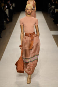 Model walks the runway in a peach short-sleeve top and pink patterned skirt with a wide woven belt, carrying an orange knit bag.