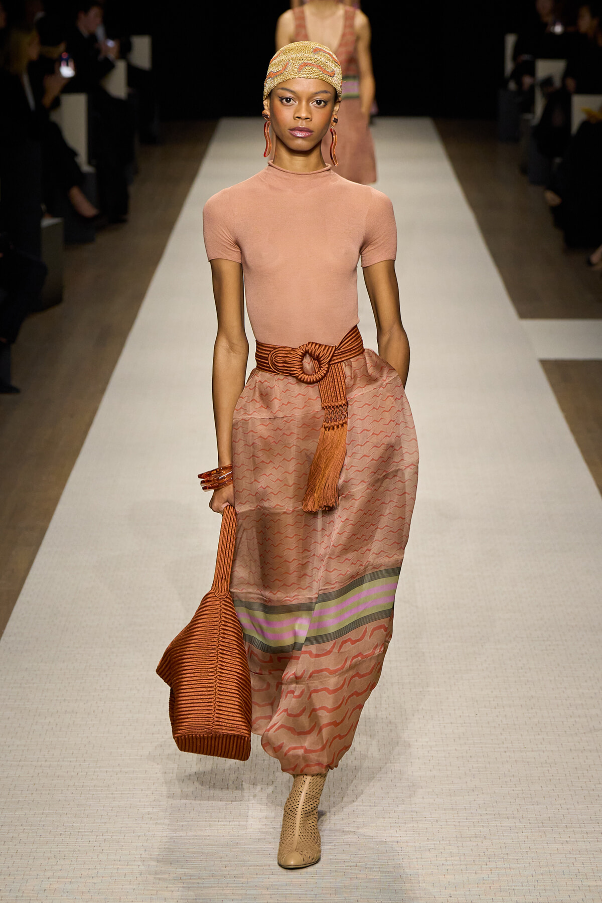 Model walks the runway in a peach short-sleeve top and pink patterned skirt with a wide woven belt, carrying an orange knit bag.