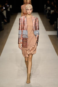 Model walking the runway in a peach-toned satin dress with a patterned blazer, beige head wrap, and matching boots on a white catwalk.