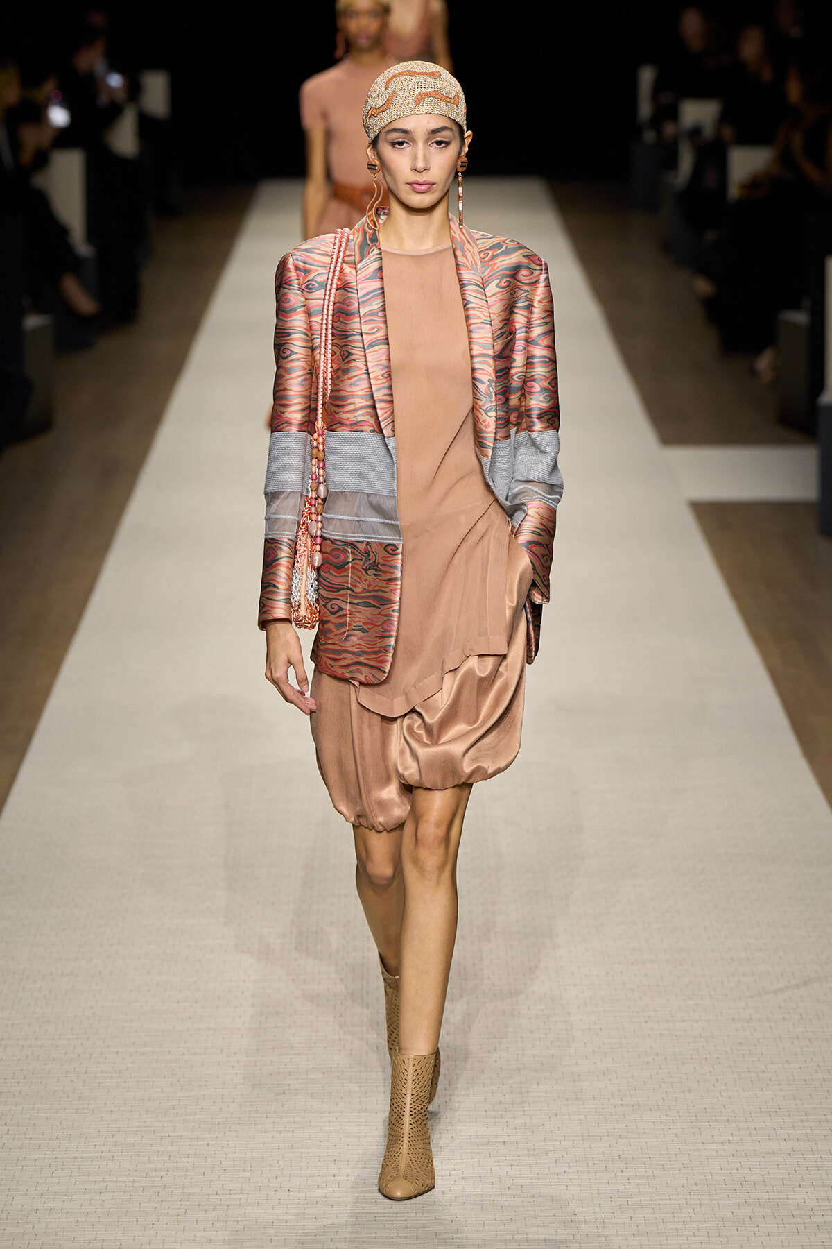 Model walking the runway in a peach-toned satin dress with a patterned blazer, beige head wrap, and matching boots on a white catwalk.