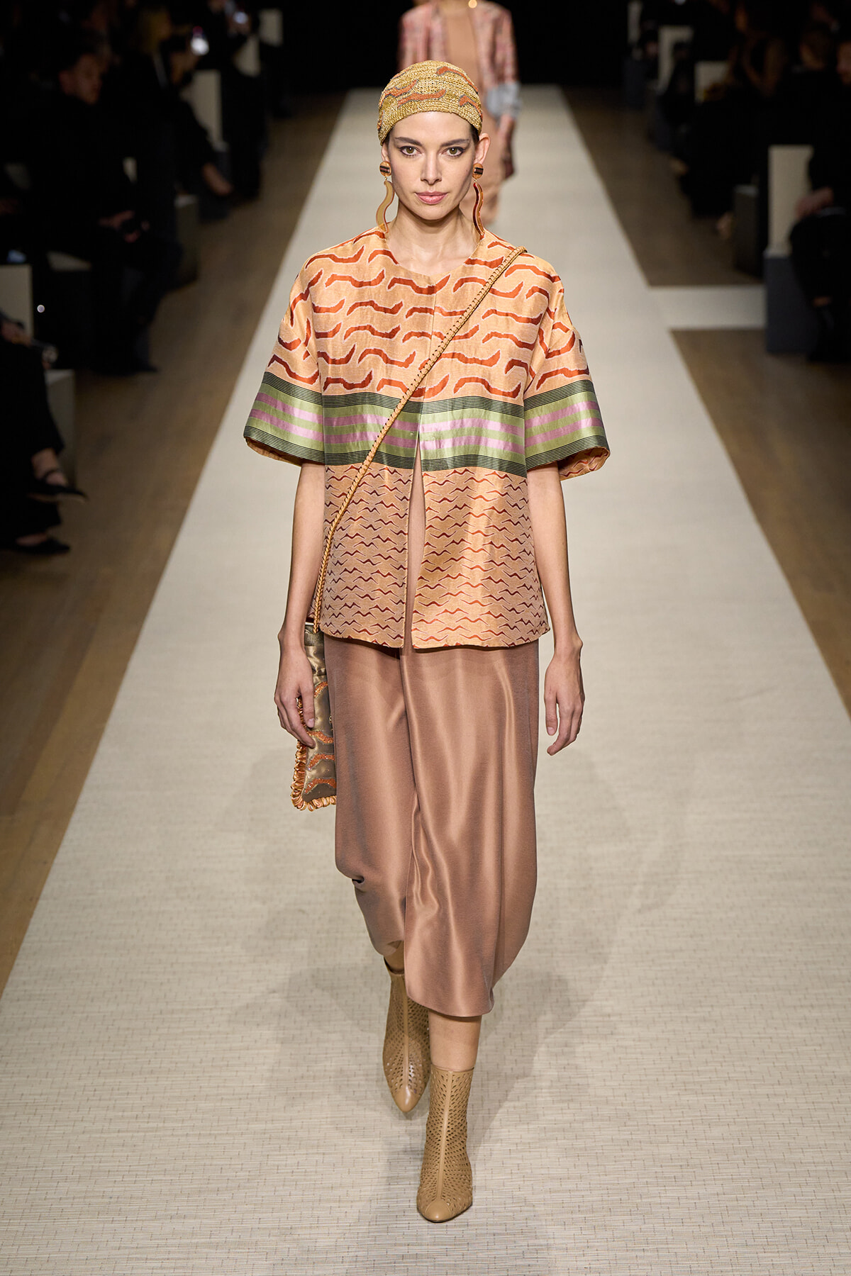 Model on runway wearing a peach patterned tunic with green and pink striped hem, satin peach skirt, tan ankle boots, and a patterned headscarf.