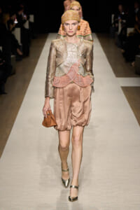 Runway model in a metallic pleated gold jacket with pink scalloped peplum, satin bronze shorts, gold heels, head wrap, and orange clutch.