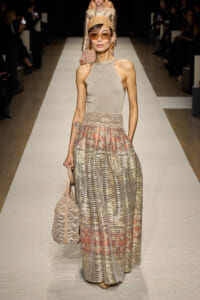 Runway model in a beige halter top and shimmering, patterned floor-length skirt with a lace waistband, headscarf, and oversized sunglasses.