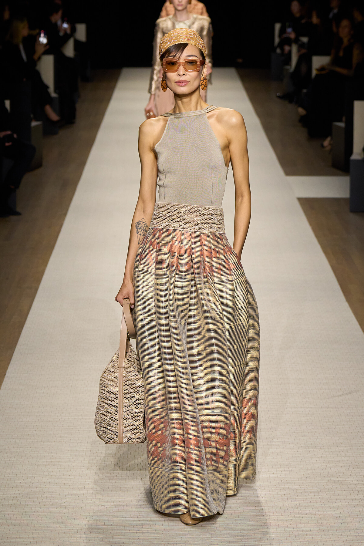 Runway model in a beige halter top and shimmering, patterned floor-length skirt with a lace waistband, headscarf, and oversized sunglasses.