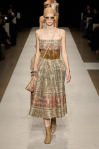 Model on a runway wearing a metallic pleated strapless dress with a wide brown belt, head scarf, oversized sunglasses, and ankle boots.