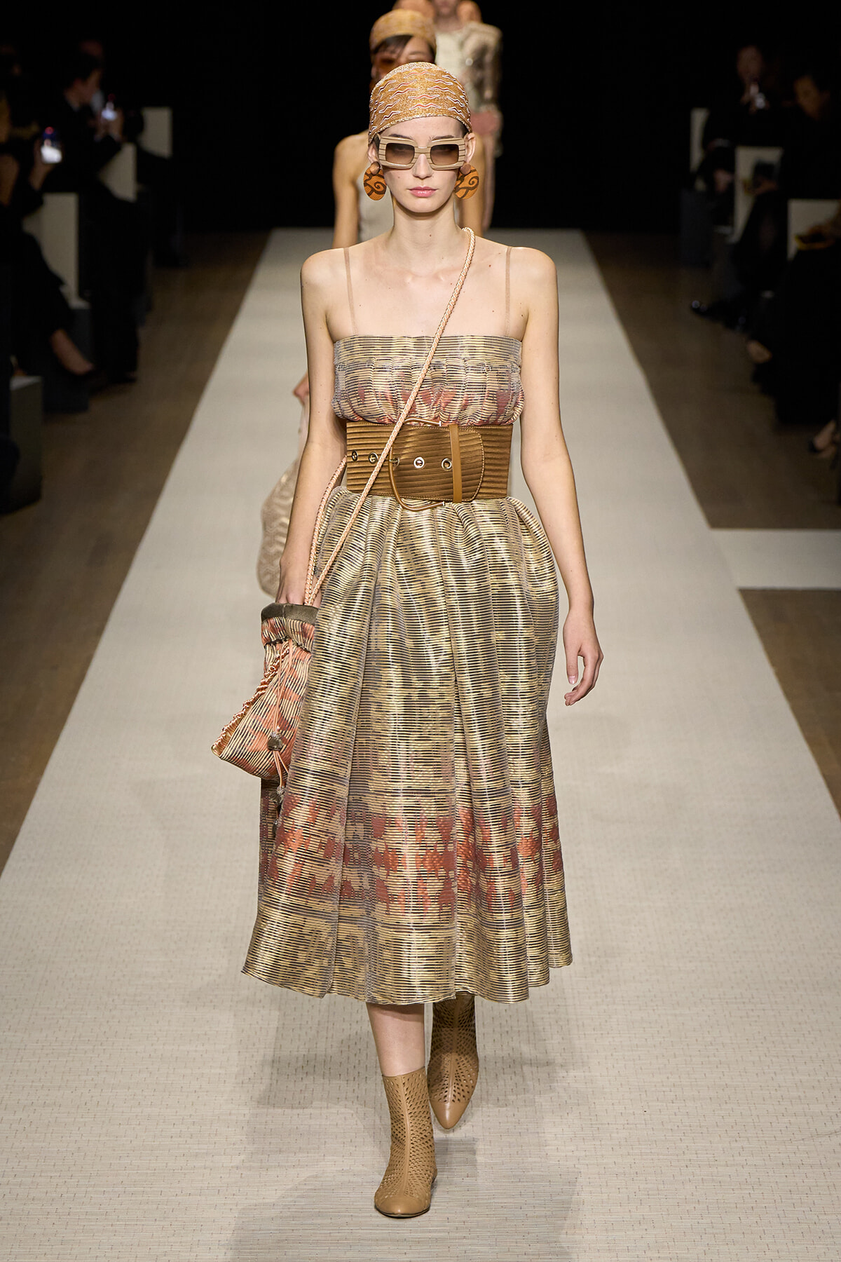 Model on a runway wearing a metallic pleated strapless dress with a wide brown belt, head scarf, oversized sunglasses, and ankle boots.