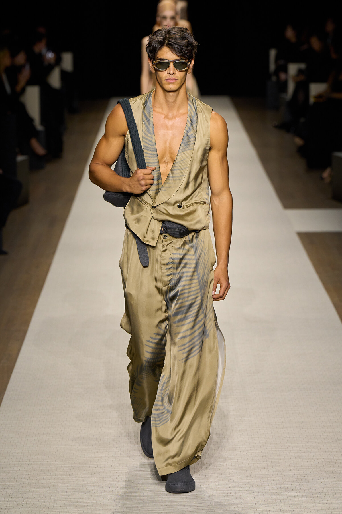 Male model on a runway wearing a tan satin sleeveless jumpsuit with blue tie-dye pattern, sunglasses, and a black shoulder bag.
