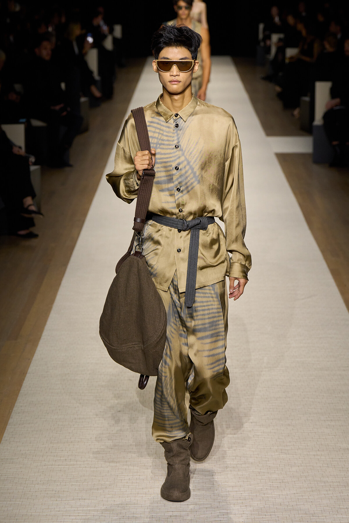 Male model walks the runway in a tan satin shirt and matching pants with a blue tie-dye pattern, carrying a brown shoulder bag and wearing oversized sunglasses.