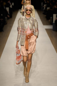Model walking a runway in a peach satin bubble-hem shorts, metallic striped jacket, headscarf, chunky sunglasses, and tan ankle boots, carrying a woven peach bag