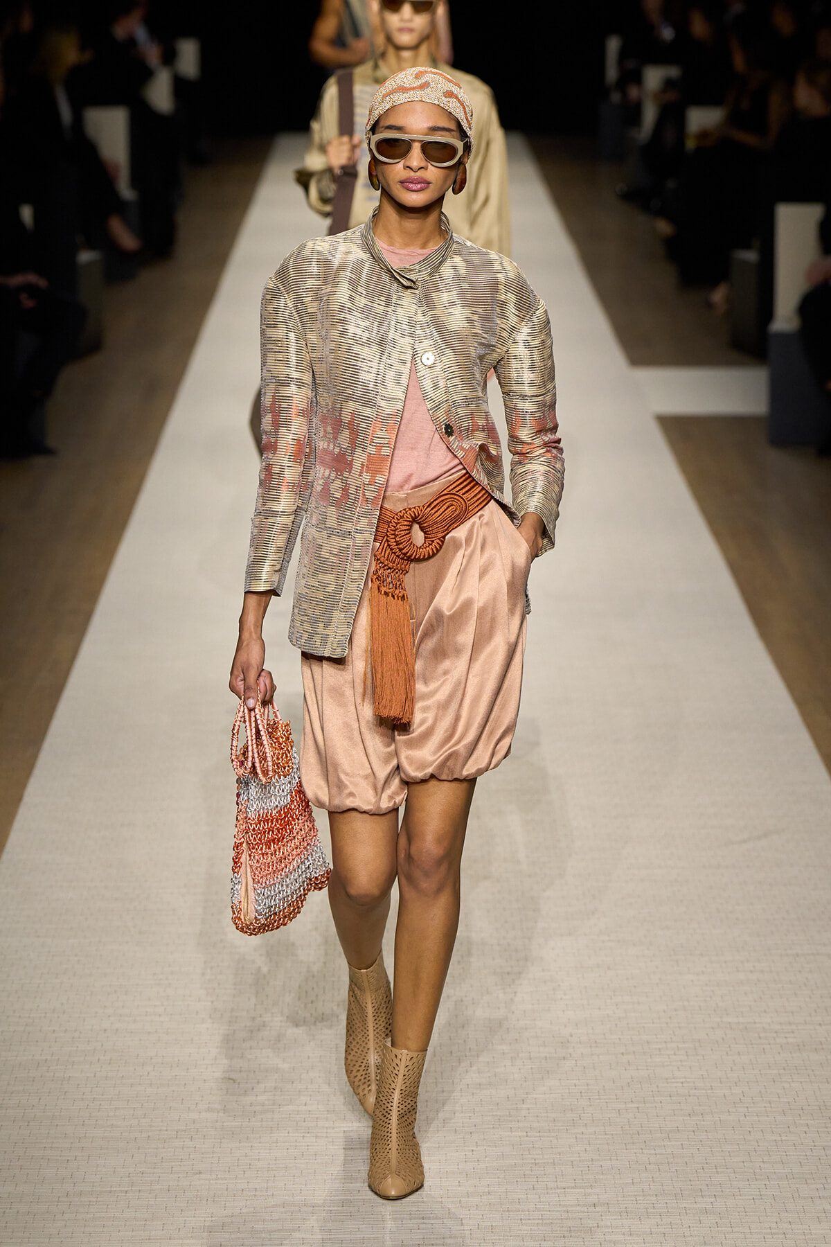 Model walking a runway in a peach satin bubble-hem shorts, metallic striped jacket, headscarf, chunky sunglasses, and tan ankle boots, carrying a woven peach bag