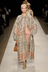 Model walks a runway in a long, patterned coat with a peach underlayer, headscarf, and tan ankle boots, carrying a brown bag.