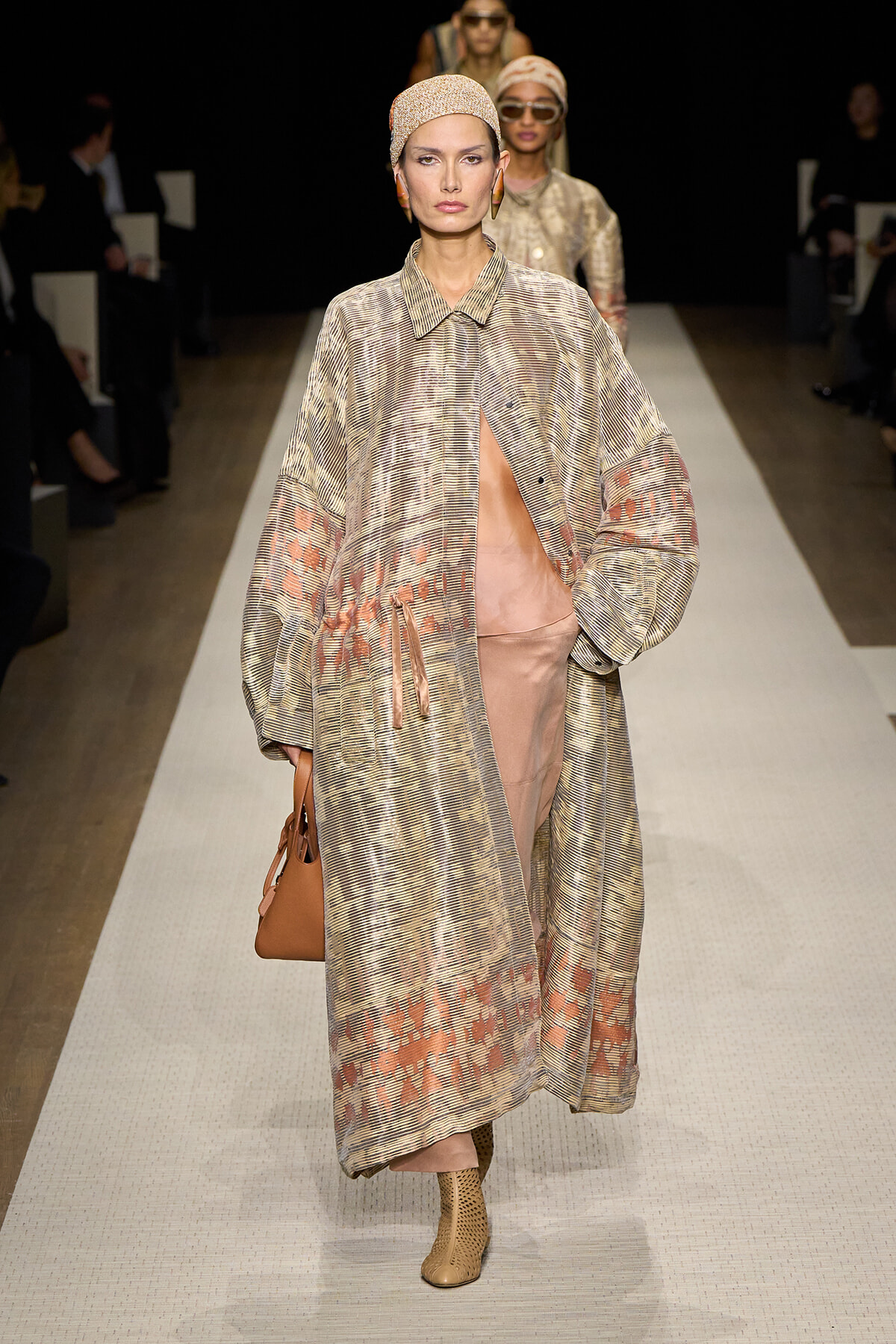 Model walks a runway in a long, patterned coat with a peach underlayer, headscarf, and tan ankle boots, carrying a brown bag.
