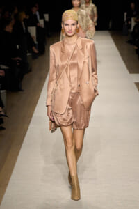 Model on a runway wearing a peach satin blazer with matching short satin shorts, headscarf, statement earrings, tan ankle boots, and a small fringed crossbody bag.