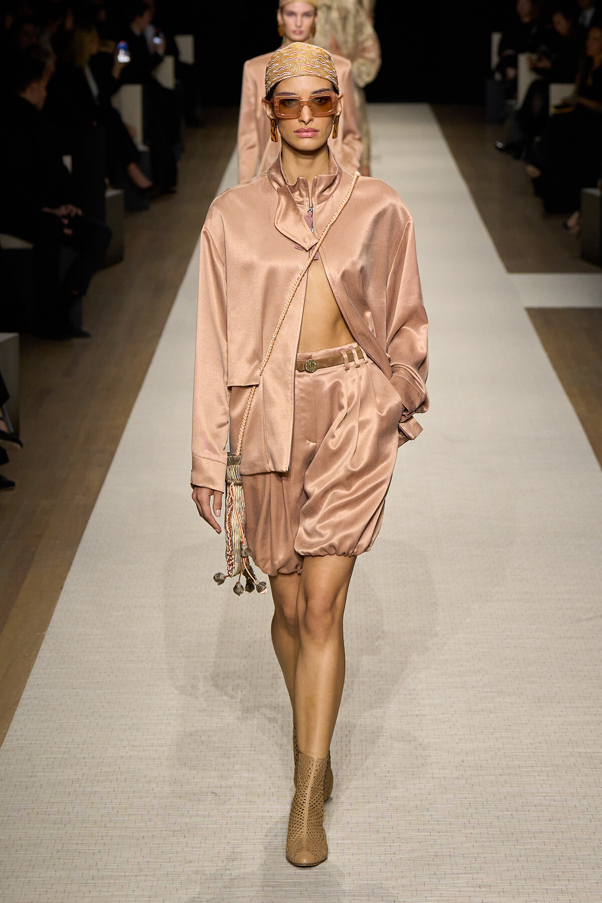 Model in pale pink satin suit walks the runway with a draped jacket, bubble shorts, headscarf, sunglasses, and a crossbody chain bag.