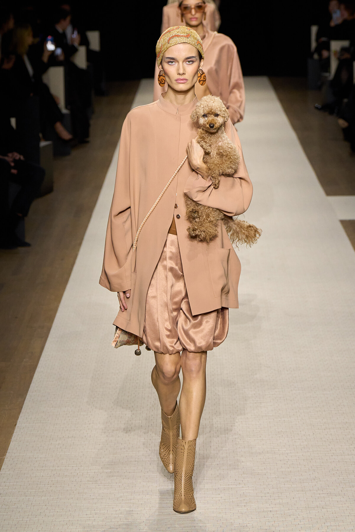 Model walks a peach-toned ensemble down the runway, holding a small fluffy dog in a pink-beige coat and wearing a patterned headscarf and oversized earrings.