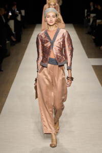 Model walks the runway in a copper satin suit: metallic embroidered jacket over a matching top and loose wide-leg pants, headscarf, and stacked bracelets.