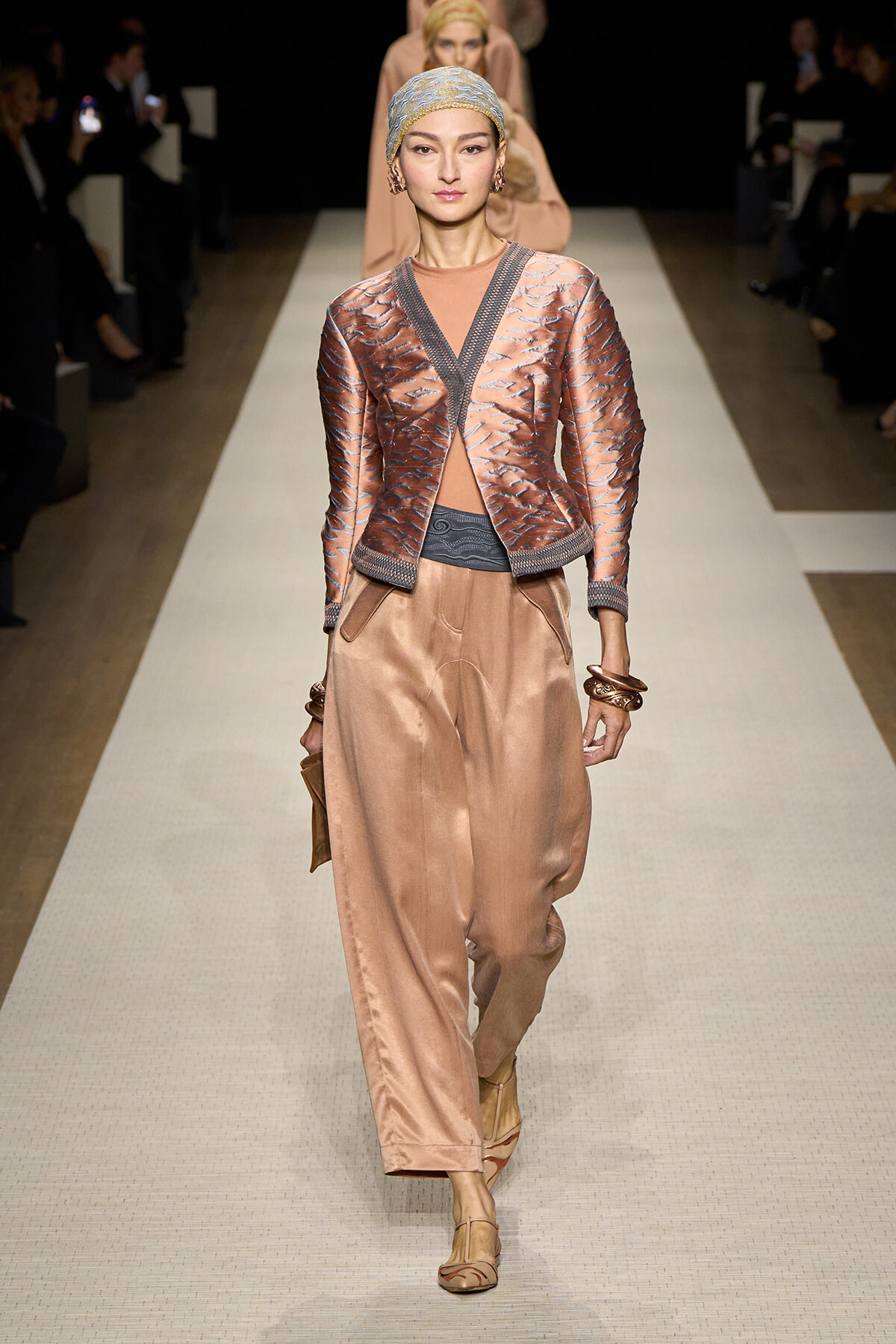Model walks the runway in a copper satin suit: metallic embroidered jacket over a matching top and loose wide-leg pants, headscarf, and stacked bracelets.
