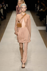 Model walking a fashion runway in a peach-toned asymmetric outfit with a matching headscarf and a small tan crossbody bag across the chest.