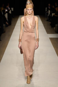 Model walking a fashion runway in a satin taupe gown with a deep V neckline, headscarf, chunky bracelets, and ankle boots.