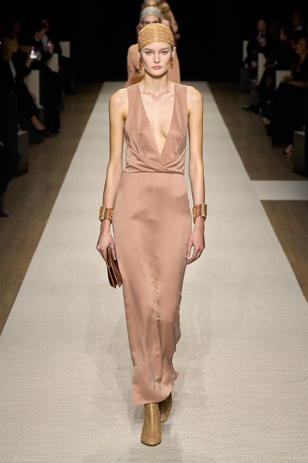 Model walking a fashion runway in a satin taupe gown with a deep V neckline, headscarf, chunky bracelets, and ankle boots.