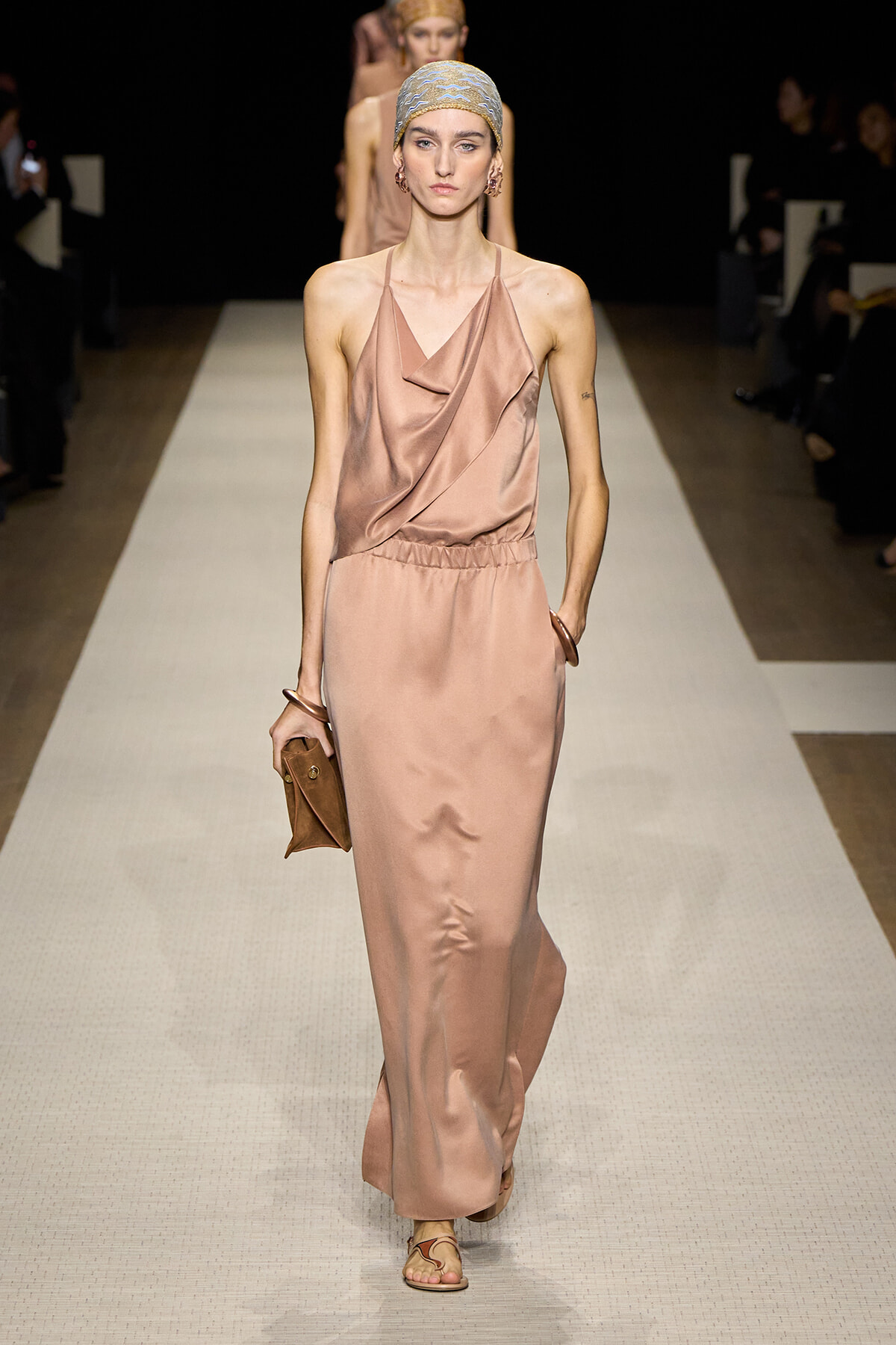 Model walking a fashion runway in a blush satin draped halter dress, headscarf, matching sandals, and a small brown clutch.
