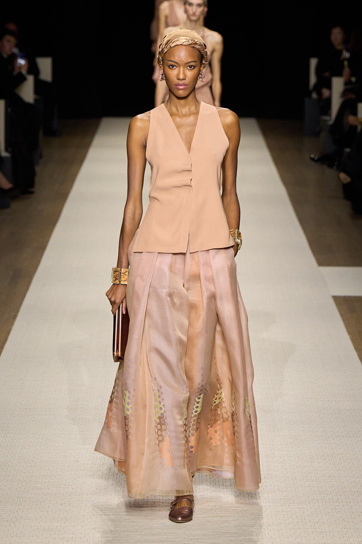 Model walks the runway in a peach sleeveless V-neck top paired with a flowing, peach-toned skirt featuring sheer color-block panels and metallic embroidery, headwrap and gold jewelry.