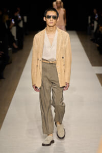 Male model walks the runway in a beige blazer, light striped shirt, braided belt, olive trousers, and tan loafers with sunglasses.