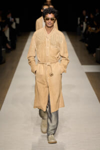 Male model on a runway wearing a beige belted knee-length coat and sunglasses.