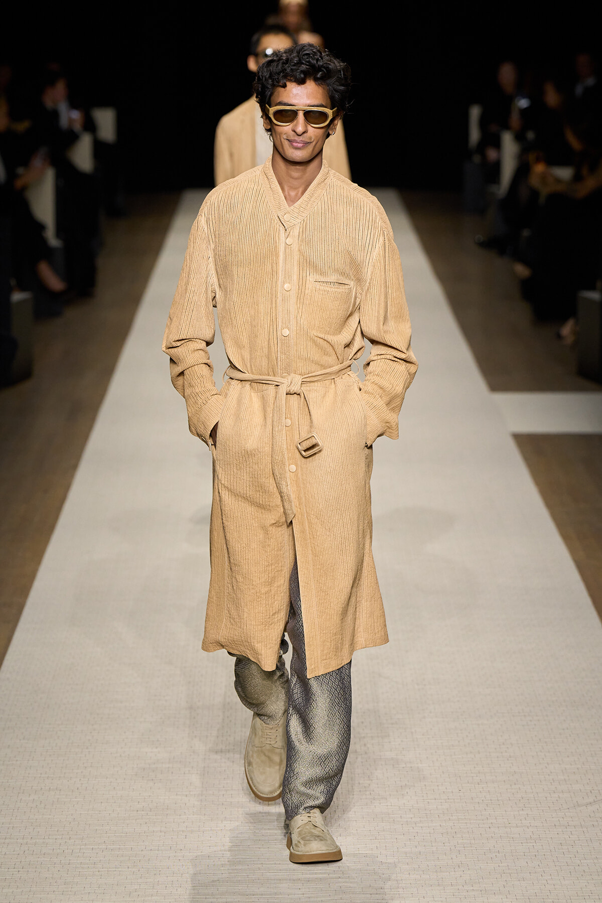 Male model on a runway wearing a beige belted knee-length coat and sunglasses.