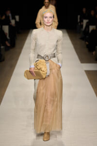 Runway model in a beige knit buttoned top with a wide belt and sheer tan skirt, tan boots, yellow headscarf, and tan clutch.