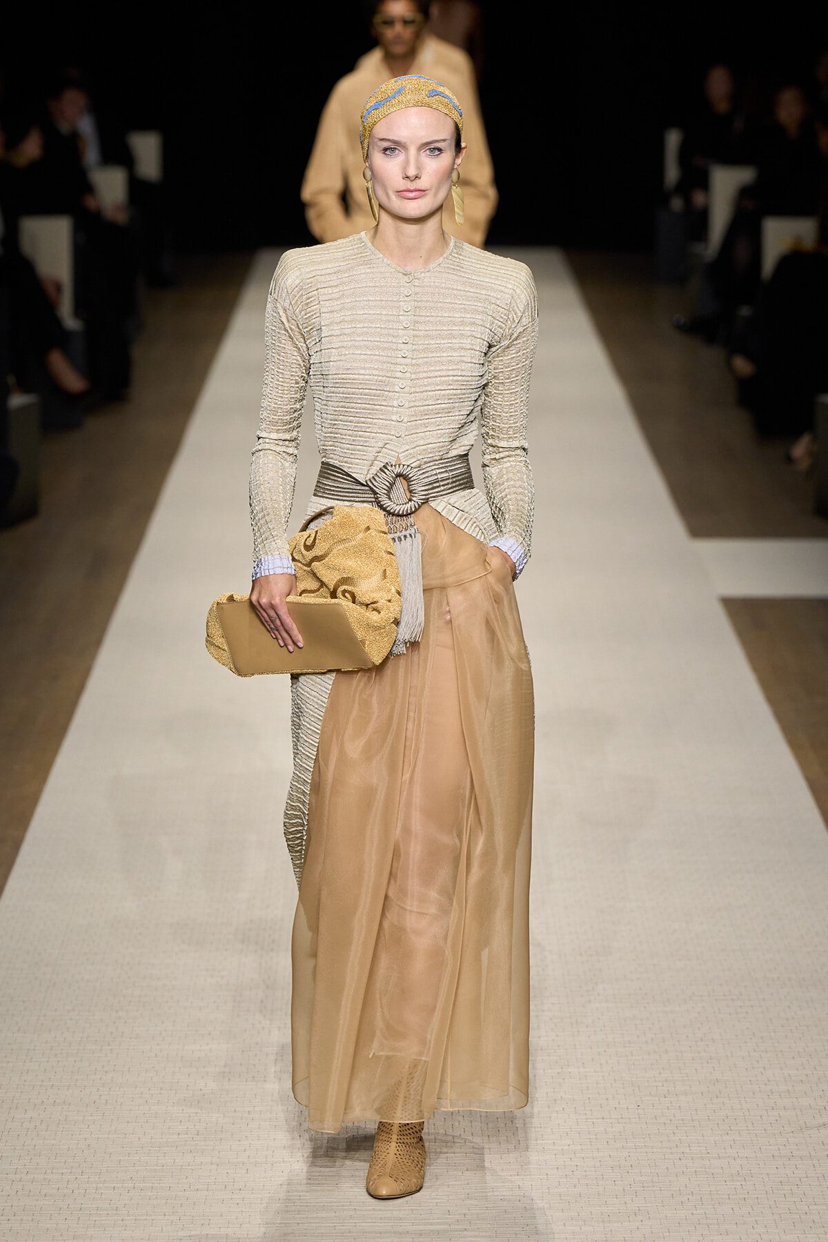 Runway model in a beige knit buttoned top with a wide belt and sheer tan skirt, tan boots, yellow headscarf, and tan clutch.