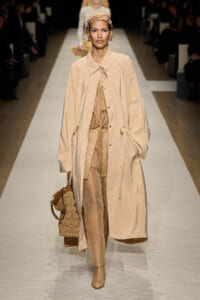 Runway model in a tan long trench over a beige matching outfit with a woven handbag.