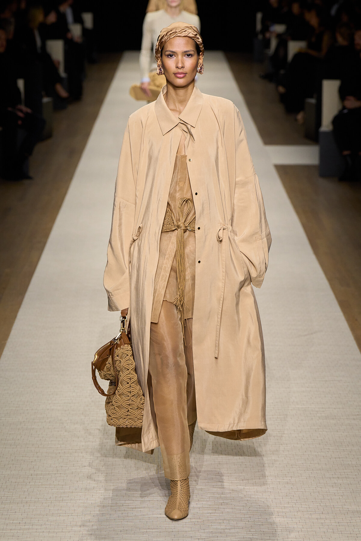 Runway model in a tan long trench over a beige matching outfit with a woven handbag.