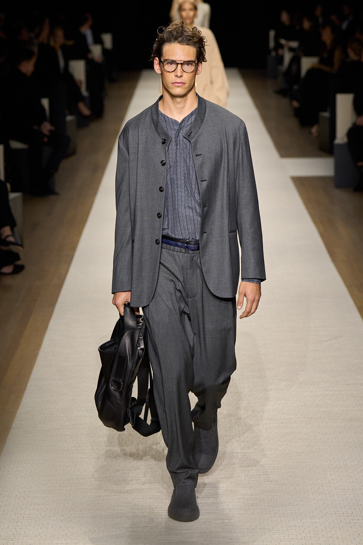 Male model walking a fashion runway in a gray blazer and matching trousers, carrying a black leather bag and wearing glasses.