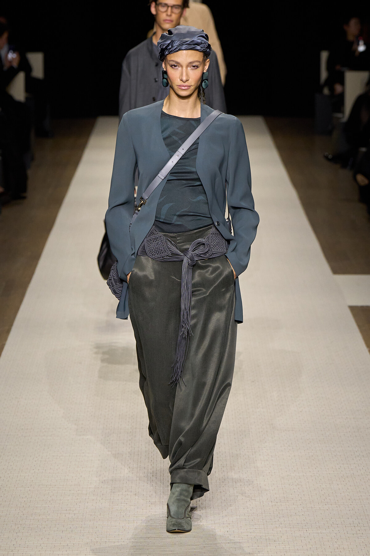 Model walking a fashion runway in a blue-gray blazer over a dark teal top, with a wide knotted belt and matching head wrap.