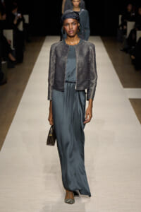 Female model walks a fashion runway in a teal satin jumpsuit with a metallic cropped jacket, black headscarf, hoop earrings, and beige heeled sandals, carrying a small brown handbag.