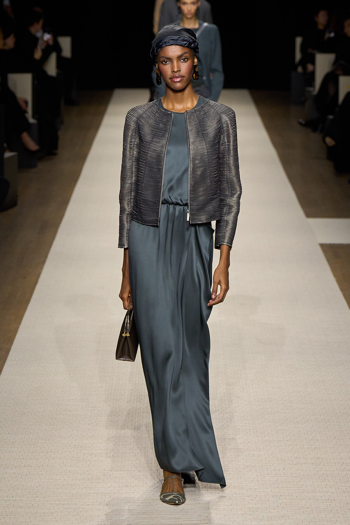 Female model walks a fashion runway in a teal satin jumpsuit with a metallic cropped jacket, black headscarf, hoop earrings, and beige heeled sandals, carrying a small brown handbag.