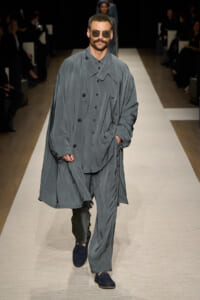 Male model walking a fashion runway in an oversized gray trench coat over a matching gray suit, wearing sunglasses and with hands in pockets.