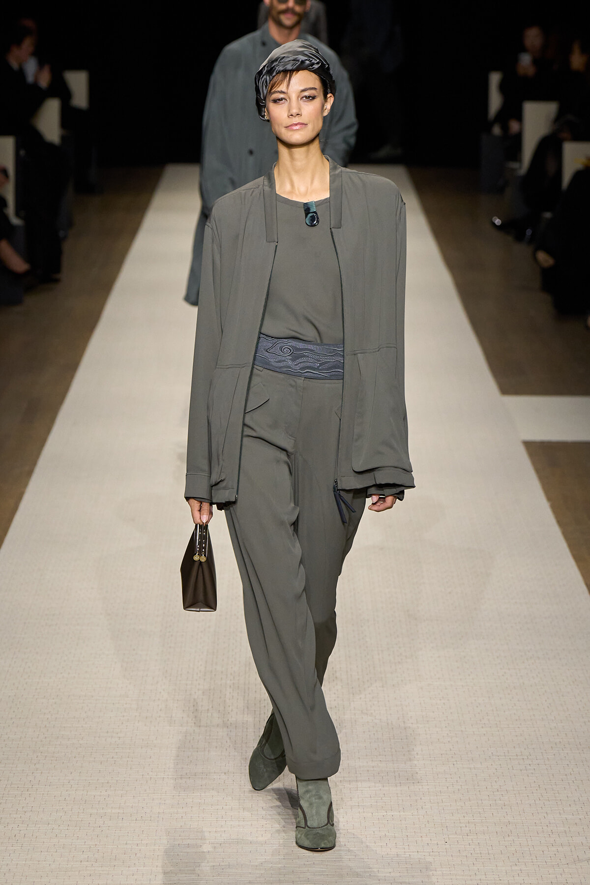 Model walking a grey-toned runway wearing a relaxed matching suit with a head scarf and belt, carrying a small brown handbag.