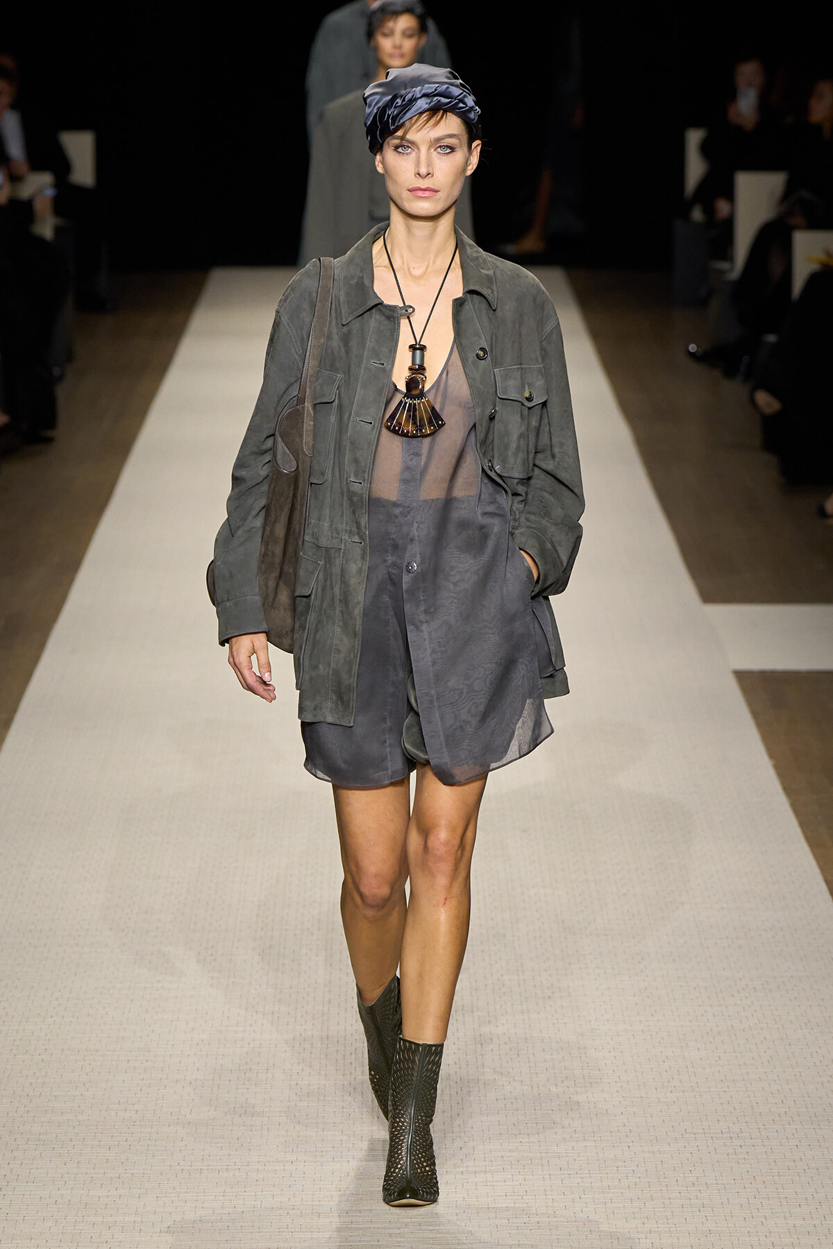 Model walks a gray layered outfit on a runway: oversized jacket, sheer top, matching shorts, ankle boots, and a satin turban-style hat with a bold necklace kept open at the neck.