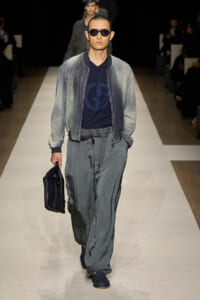 Male model walks a fashion runway in a grey gradient bomber jacket, navy logo tee, loose grey pants, black belt, sunglasses, and a black briefcase.