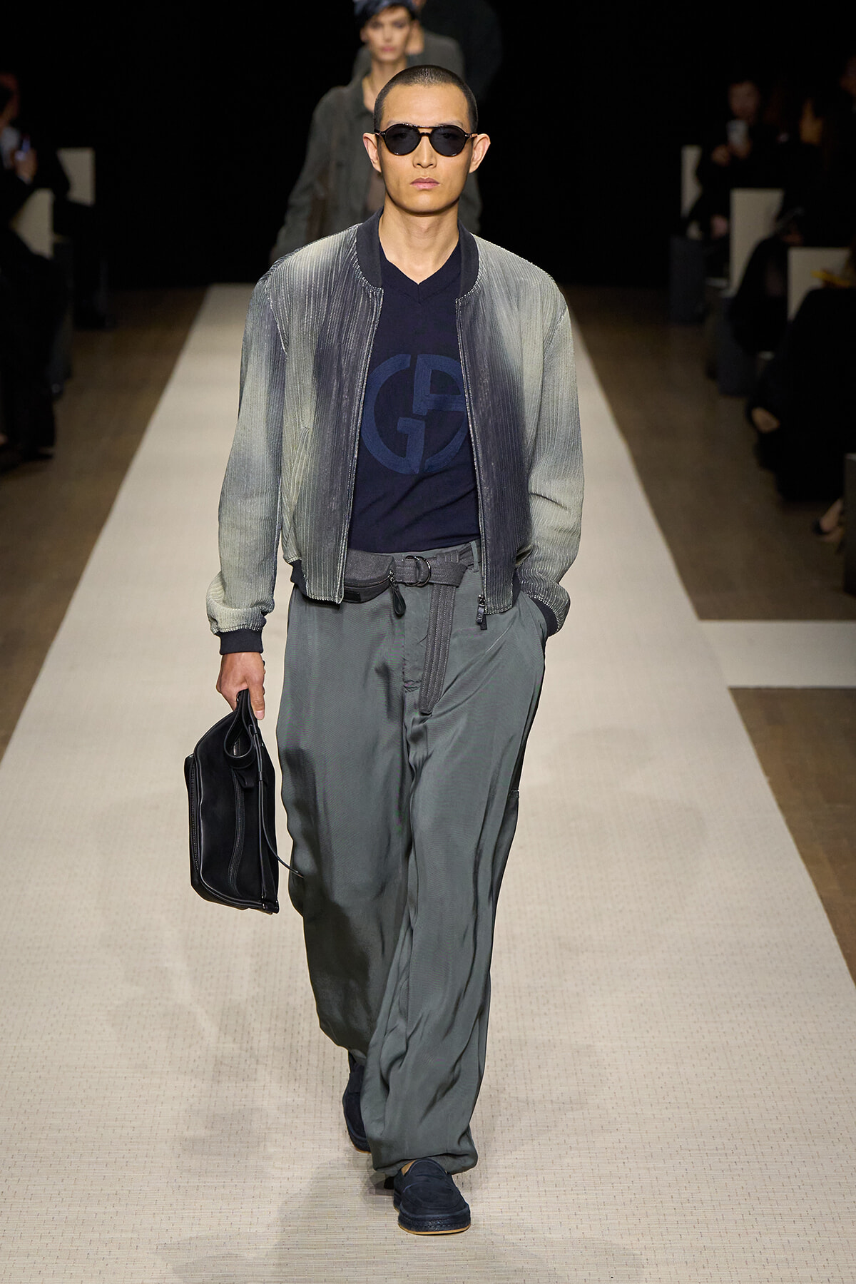 Male model walks a fashion runway in a grey gradient bomber jacket, navy logo tee, loose grey pants, black belt, sunglasses, and a black briefcase.