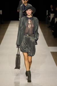 Model on a runway wearing a sheer black top with front ties, an oversized olive-green jacket, a gray satin bubble skirt, a wide-brim hat, and black ankle boots, carrying a gray handbag.